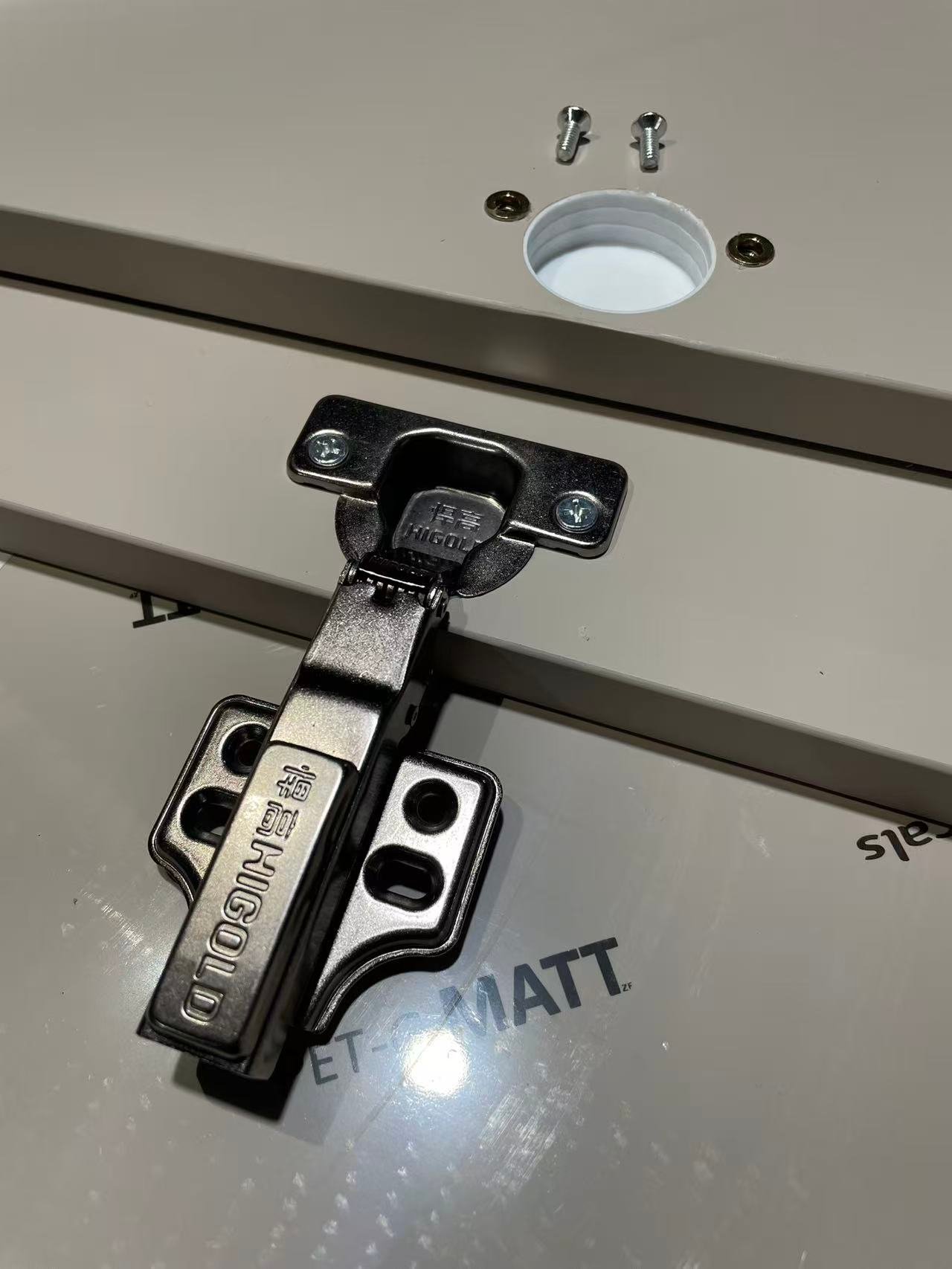 Concealed hinge test-fitted on an aluminum cabinet door beside its drilled cup hole and screws